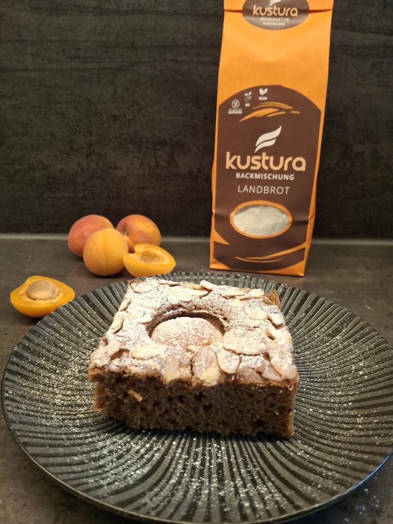 glutenfreier Marillenkuchen mit KUSTURA Landbrot‑Backmischung, goldbraun gebacken, belegt mit frischen Marillenhälften und Mandelblättern, serviert auf einem dunklen Teller vor der KUSTURA‑Verpackung